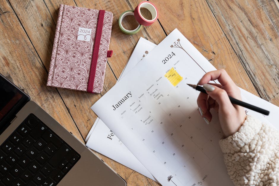 A person plans events in a 2024 calendar beside a notebook and laptop on a wooden desk.